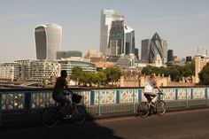 two people riding bikes on a bridge in front of the city