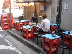 two people sitting at tables with red crates on the side of the street in front of them