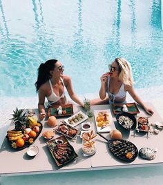 two women sitting at a table with food near a pool