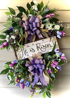 a wreath with purple and white flowers hanging on the side of a house that says he is risen