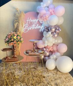 a birthday party setup with balloons and flowers on the side table in front of a sign that says happy birthday