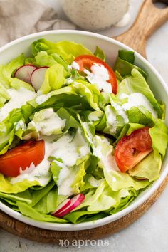 a white bowl filled with lettuce, tomatoes and dressing next to a wooden spoon