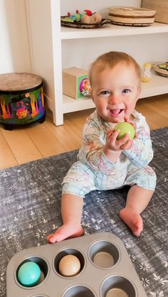 a baby is sitting on the floor eating from a muffin tin and holding an apple