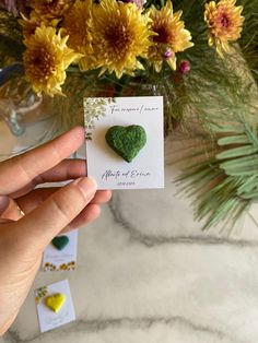 a person holding up a card with a heart shaped green stone on it next to flowers