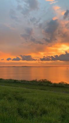 the sun is setting over the ocean with clouds in the sky and grass on the ground