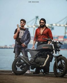 two men standing next to each other near a motorcycle on the water's edge