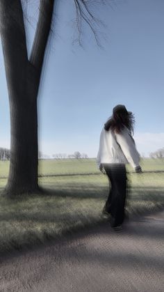 a woman walking down a road next to a tree and grass covered field with no leaves