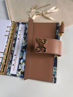 an open notebook with a gold butterfly on the front cover and ribbon attached to it