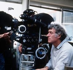 a man sitting in front of a camera on top of a wooden table next to other people