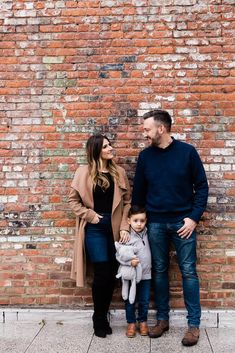 a man and woman standing in front of a brick wall with a baby on their lap