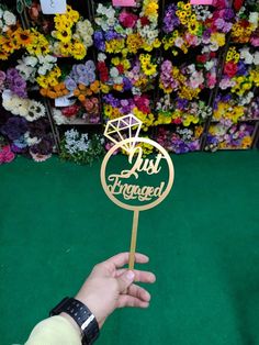 a hand holding a wooden sign that says just engaged in front of colorful flower display