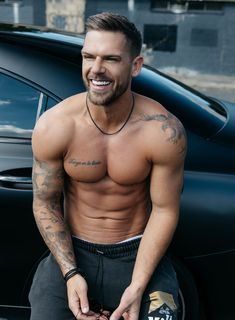 a shirtless man sitting on the hood of a car with his hands in his pockets