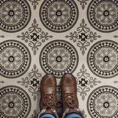 someone's feet in brown shoes and jeans standing on a patterned wallpapered floor