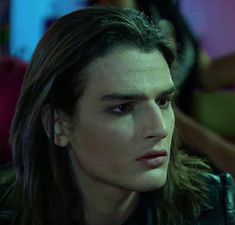 a man with long hair wearing a leather jacket and looking off to the side in front of other people