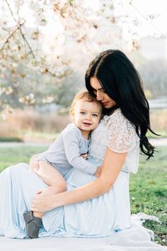 a woman holding a baby in her arms while sitting on top of a grass covered field