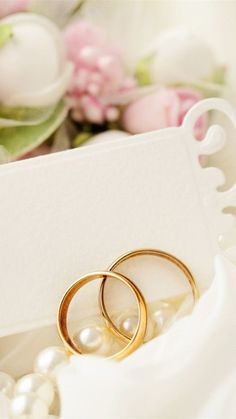 two gold wedding rings sitting on top of a white box next to pearls and flowers