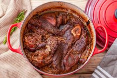 a red pot filled with meat sitting on top of a wooden table next to a cloth