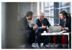 three men sitting at a table talking to each other in front of a laptop computer