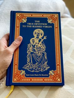 a hand holding a blue book on top of a white and yellow bed sheet with an image of the virgin mary