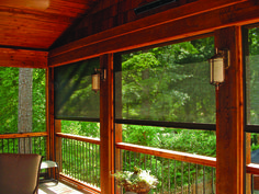 a screened porch with chairs and table on the deck overlooking wooded area in daytime light