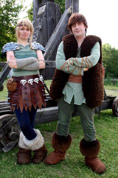 two people dressed up in costumes standing next to each other on the grass near a wooden structure