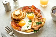 a white plate topped with eggs, bacon and pancakes next to cups of coffee on a table