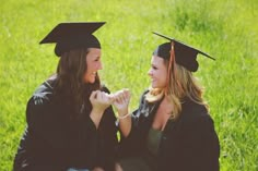 two young women in graduation gowns sitting on the grass and smiling at each other