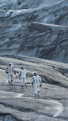 three people in white suits walking up a hill with snow on the mountains behind them