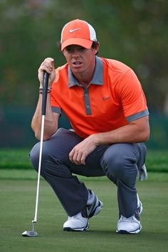 a man in an orange shirt and grey pants crouches down to put his golf ball on the green