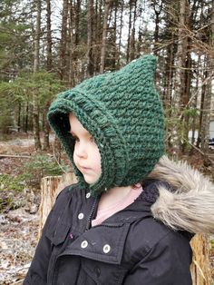 a young child wearing a green crocheted hat in the woods with trees behind it