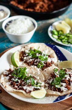 three tacos on a plate with lime wedges and other dishes in the background