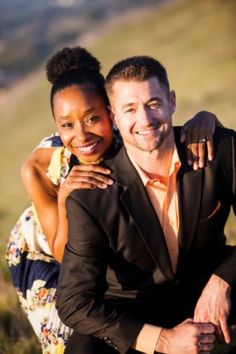 a man and woman are posing for a photo in the grass with mountains behind them