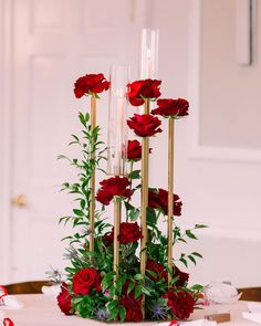 red roses and greenery are arranged in tall gold candlesticks on a table