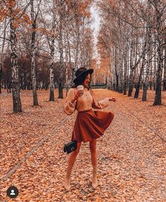 a woman in a brown dress and black hat is walking through the leaves with her cell phone