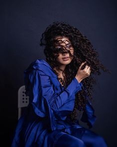 a woman with curly hair sitting on a chair wearing a blue dress and holding her hands to her face