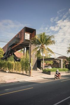 a man riding a motorcycle down a street next to a tall wooden building with a staircase on the roof