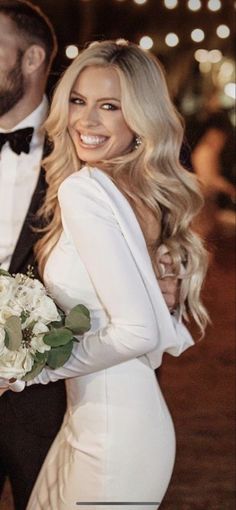 a beautiful blonde woman in a white dress holding a bouquet of flowers while standing next to a man wearing a tuxedo