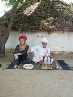 two people sitting on the ground in front of a hut