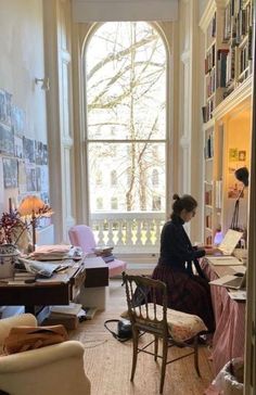 a woman sitting at a desk in front of a large window with lots of books on it