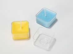 two square candles sitting next to each other on a white surface with one candle in the middle