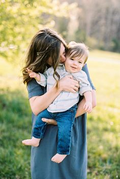 a woman holding a baby in her arms while standing on grass with trees in the background