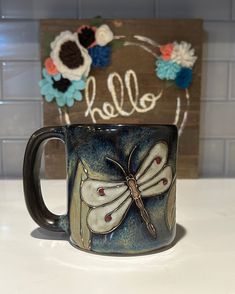 a coffee mug with a dragonfly painted on the side and hello sign behind it