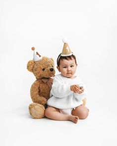 a baby sitting next to a teddy bear wearing a party hat and holding a toy