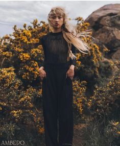 a woman with blonde hair standing in front of yellow wildflowers wearing black jumpsuit