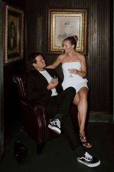 a man in a black suit and white dress sitting on a chair next to a woman