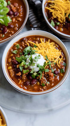 three bowls of chili with cheese and sour cream on top, sitting on a marble table