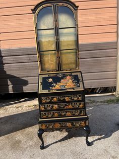 an old black and gold painted bureau desk with glass doors on the top, sitting in front of a garage door