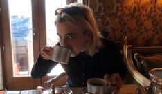 a woman sitting at a table drinking from a coffee cup and holding a mug in front of her face