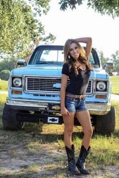 a beautiful young woman standing in front of a blue truck with her cowboy boots on