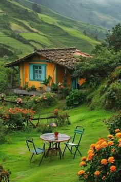 a table and chairs in front of a yellow house on a green hillside with flowers